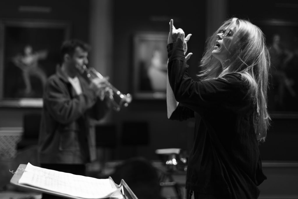 A conductor raises hands in the foreground with a trumpeter in the background, black and white