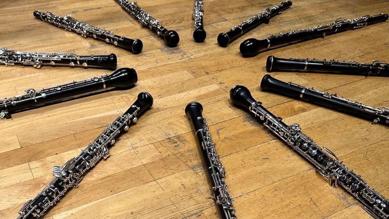 12 different types of black wind instrument with silver overlaid keywork (oboes, cor anglais etc) laid out in a clock circle on a wooden floor.
