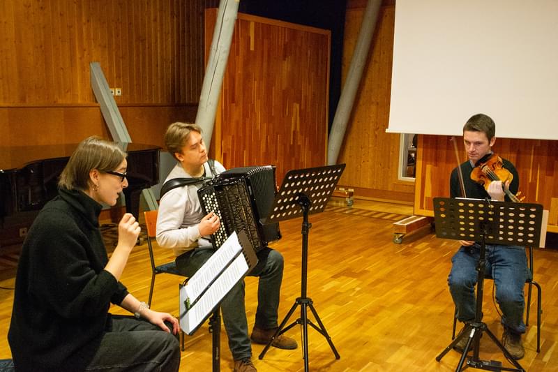 Three people sitting in a circle in a light wood panelled room, one singing, one playing a violin, one with an accordion.