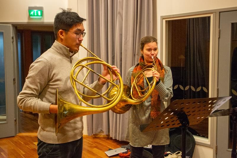 A man and a woman, each playing a curved metal horn in front of a music stand
