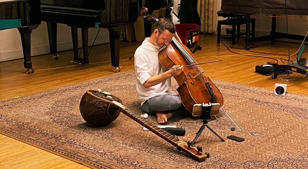 Person playing cello seated on a rug on the floor with a sitar