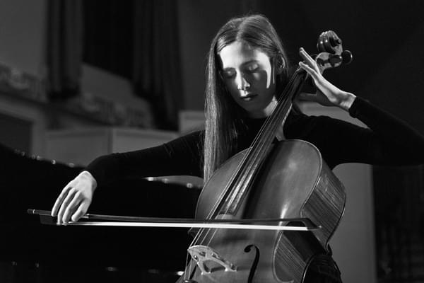 A student plays the cello alone with eyes closed in high contrast lighting (black and white)