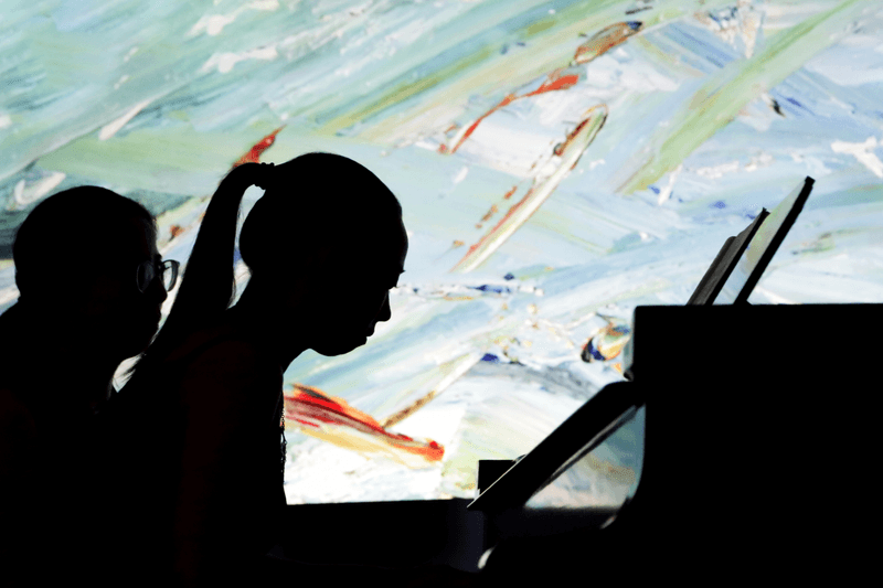 A piano student performs at a keyboard, with a mixing desk on a seat in the foreground