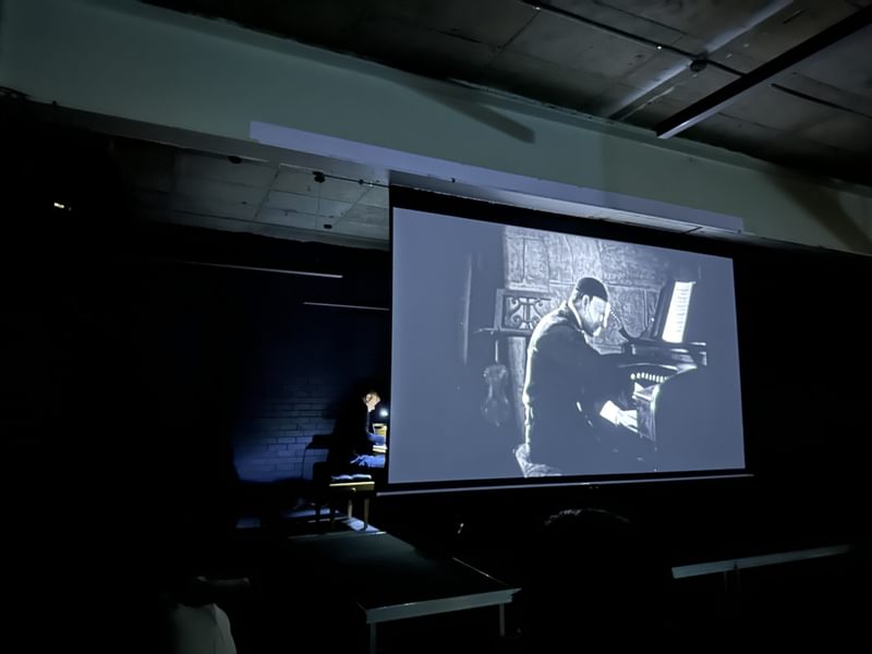 A man playing a keyboard in a dark space behind a screen showing an old black and white film of a man playing a keyboard.