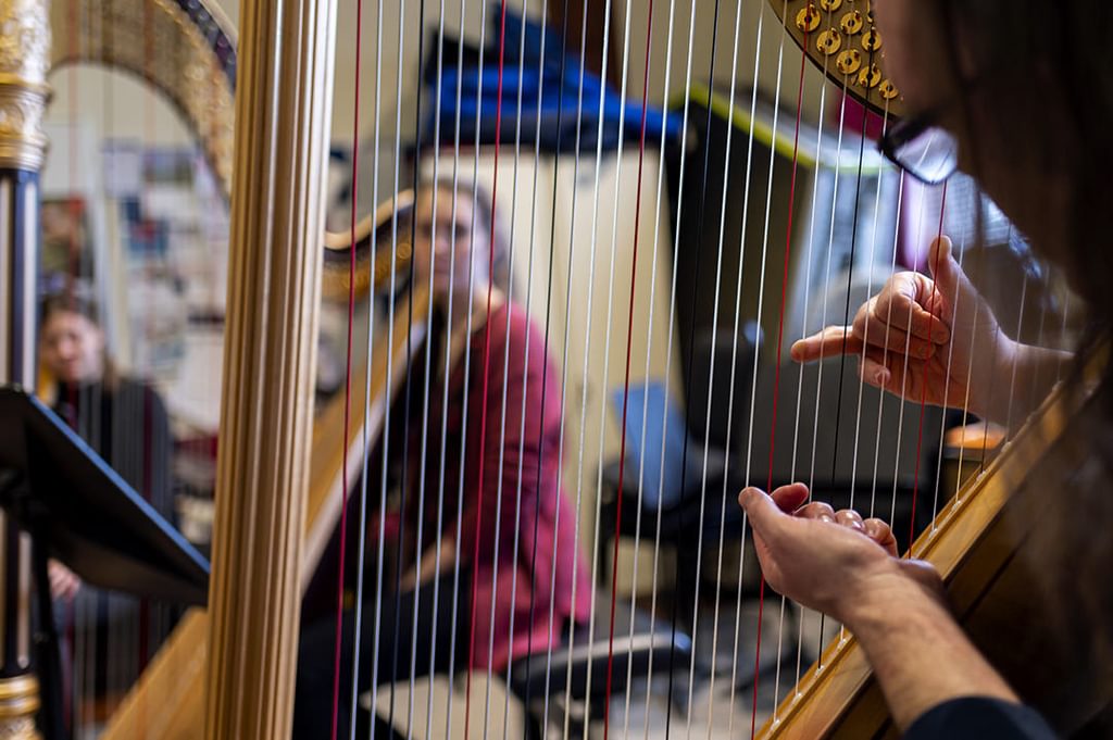A harp player's hands pluck the strings in a busy rehearsal room