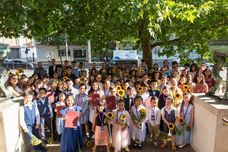 A large group of young musicians, pictured outside the Royal Academy of Music, smiling, and holding sunflowers
