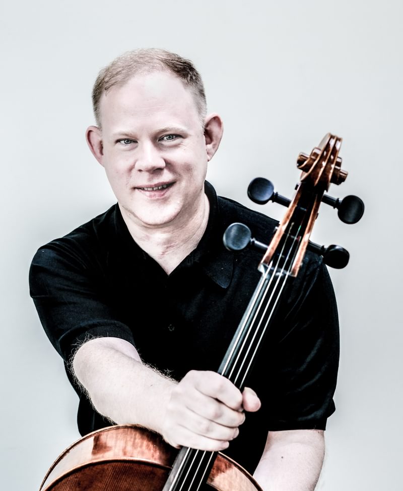 Portrait of Benjamin Hughes holding the neck of a cello, smiling for camera
