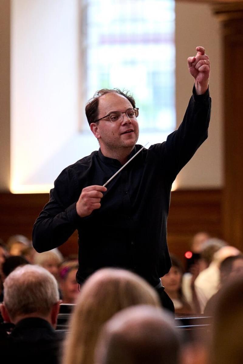 Long-distance portrait of Chris Hopkins conducting an orchestra, raising his non-baton hand