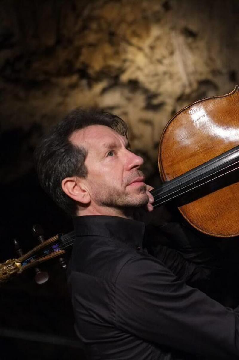 Robert Cohen holds a cello aloft over his shoulder, looking upwards